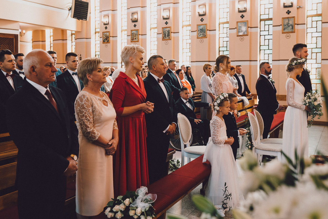 Para młoda oraz goście stoją w ławkach podczas ceremonii ślubnej w kościele.